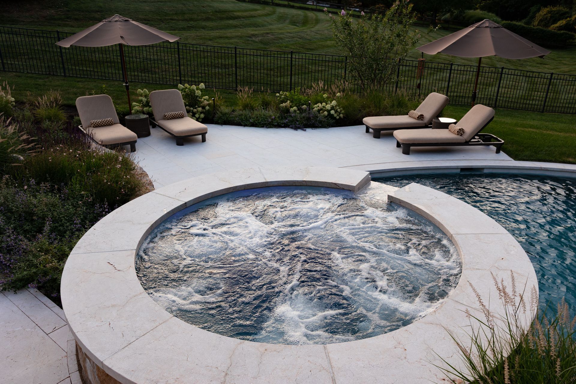 A bubbling hot tub next to a pool with lounge chairs and umbrellas on a patio.