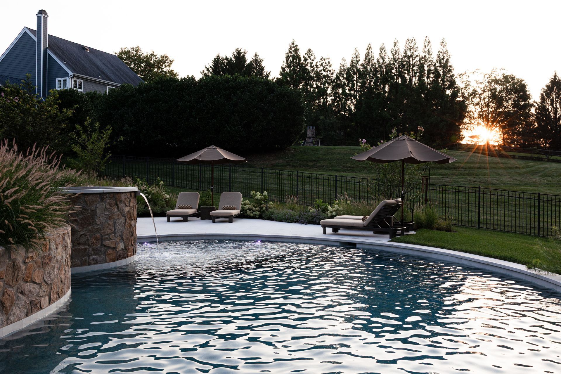Swimming pool with water feature, lounge chairs, and umbrellas in a yard at sunset.