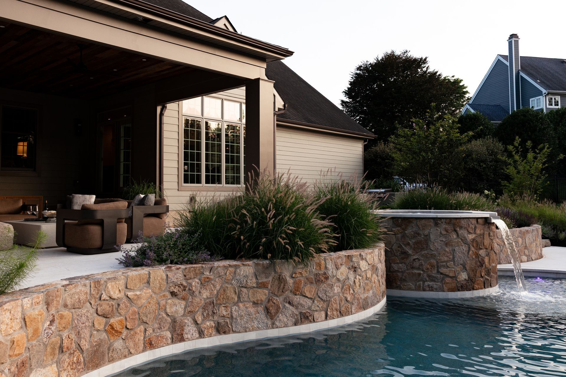 Poolside view: Stone wall with flowing water, plants, and a house with a covered patio.