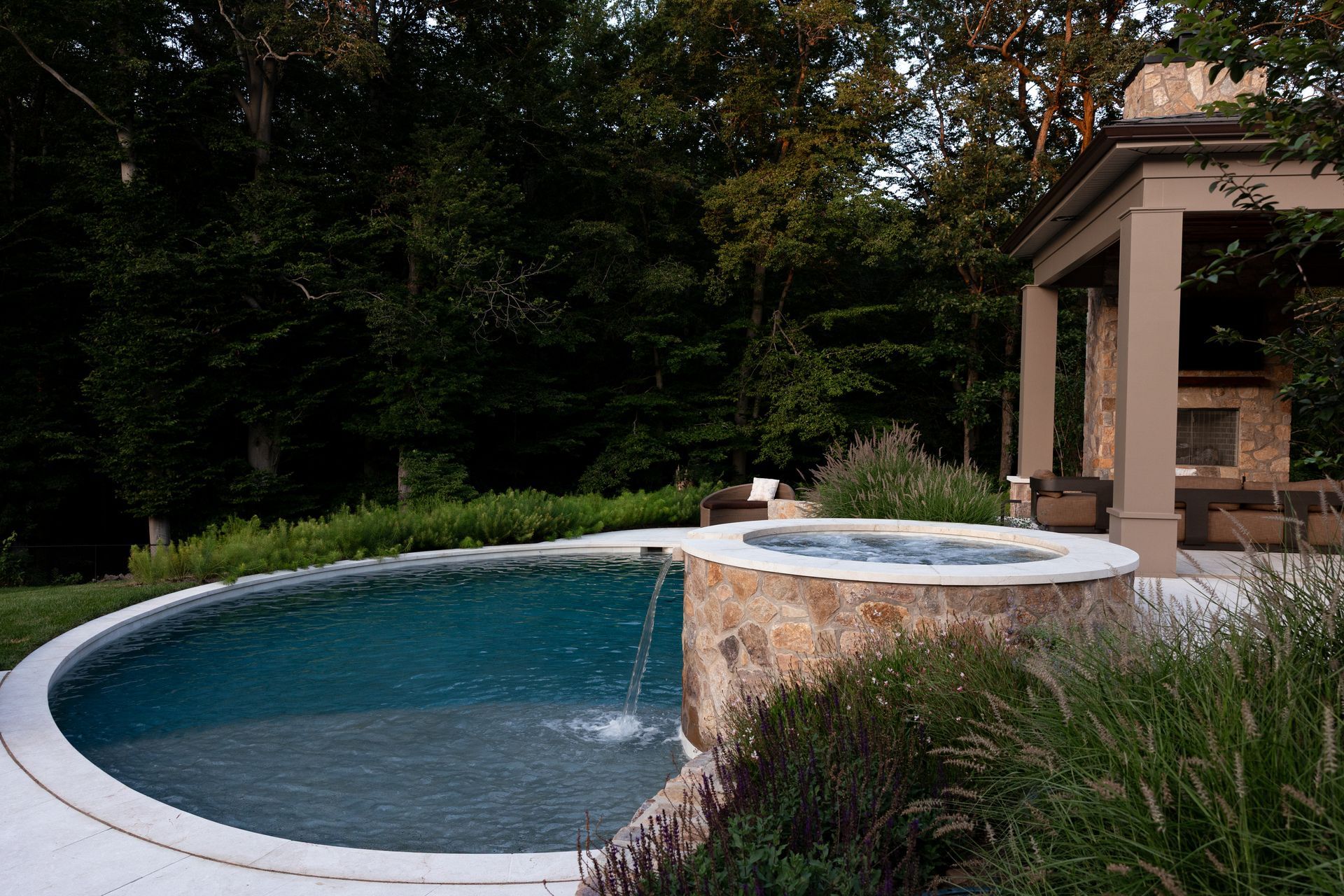 Pool with flowing water, stone hot tub, patio, and outdoor fireplace in a wooded setting.