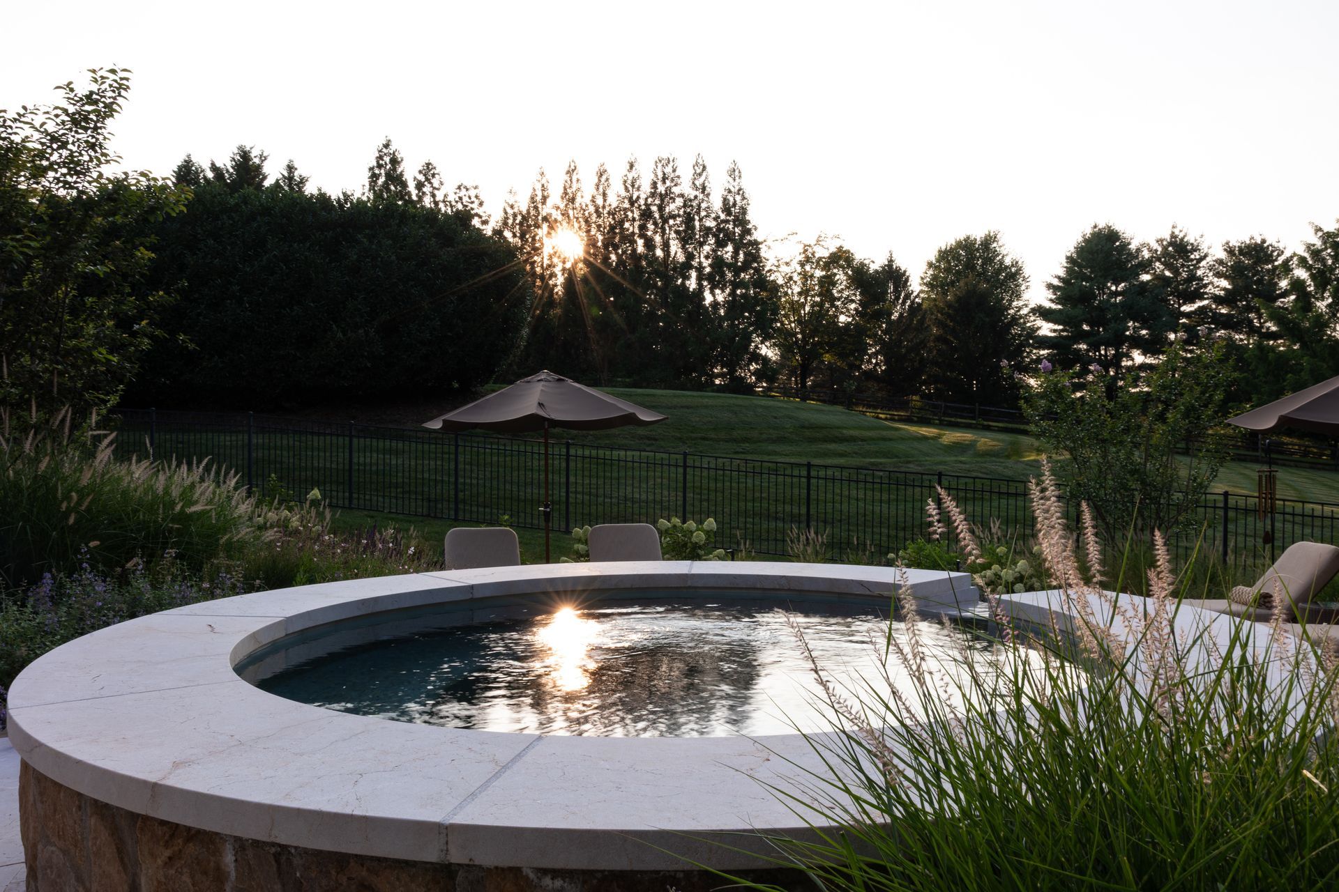 Round pool at dusk, reflecting sunlight, surrounded by stone, landscaping, and a fenced yard.