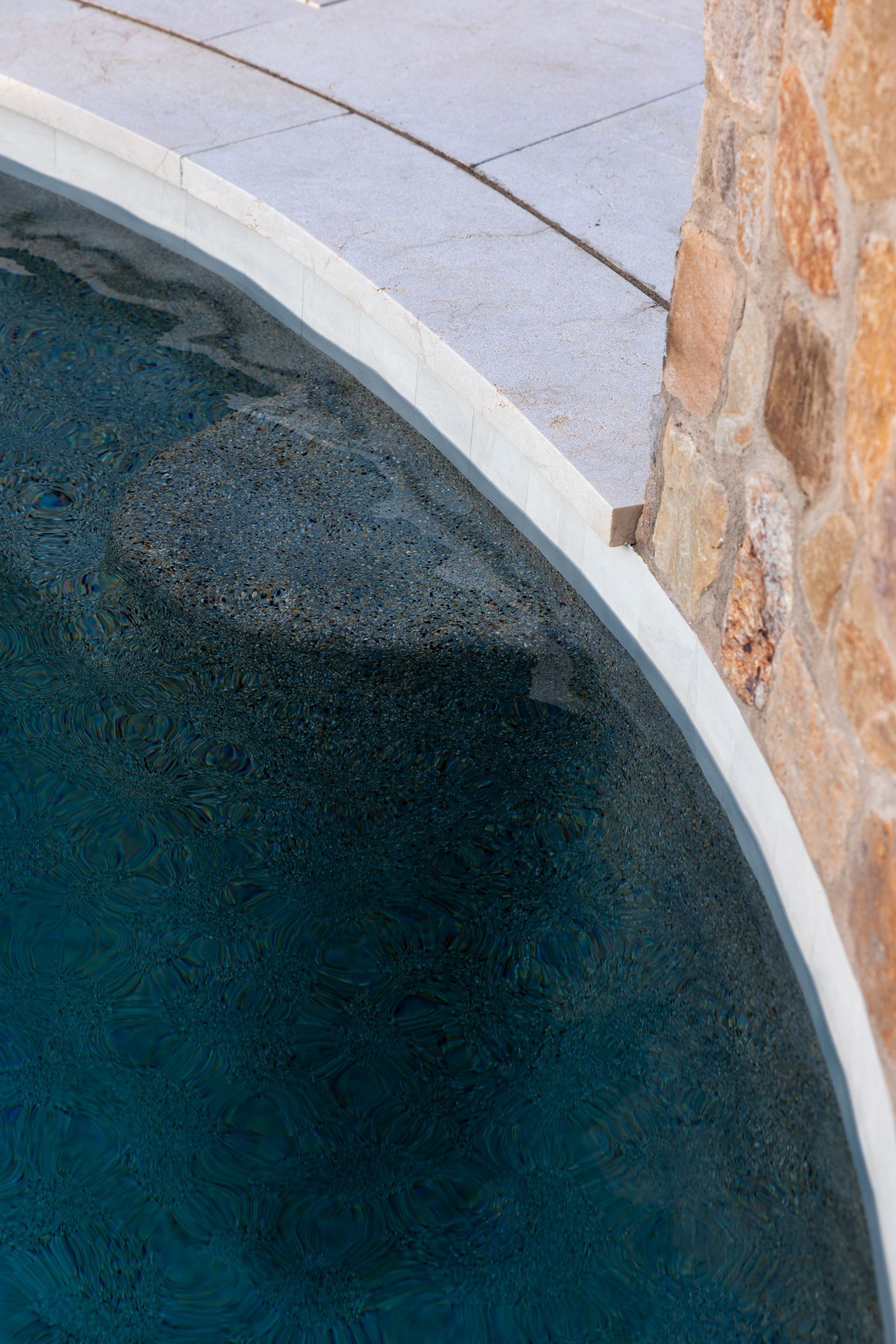 Curved pool edge with dark water next to stone wall and light colored paving.