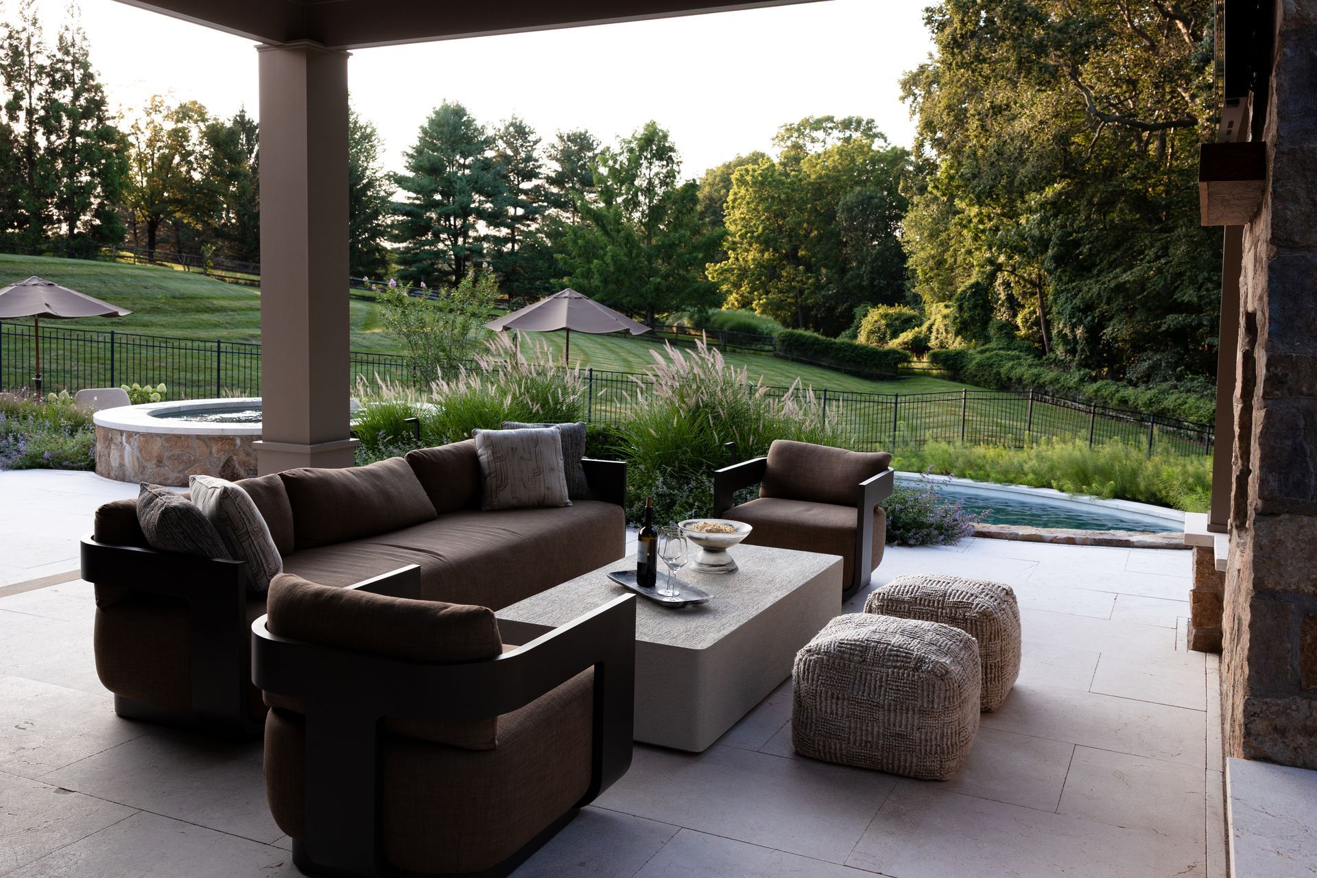 Patio with brown seating, white table, and stone ottomans overlooking a green lawn and trees.
