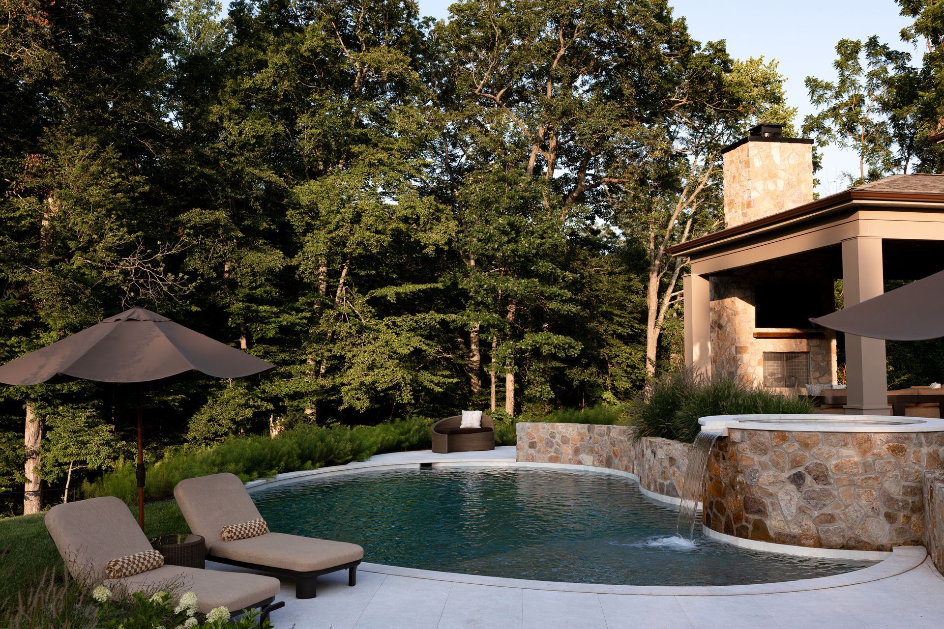 Poolside scene with lounge chairs, umbrella, spa, and stone fireplace against a backdrop of trees.