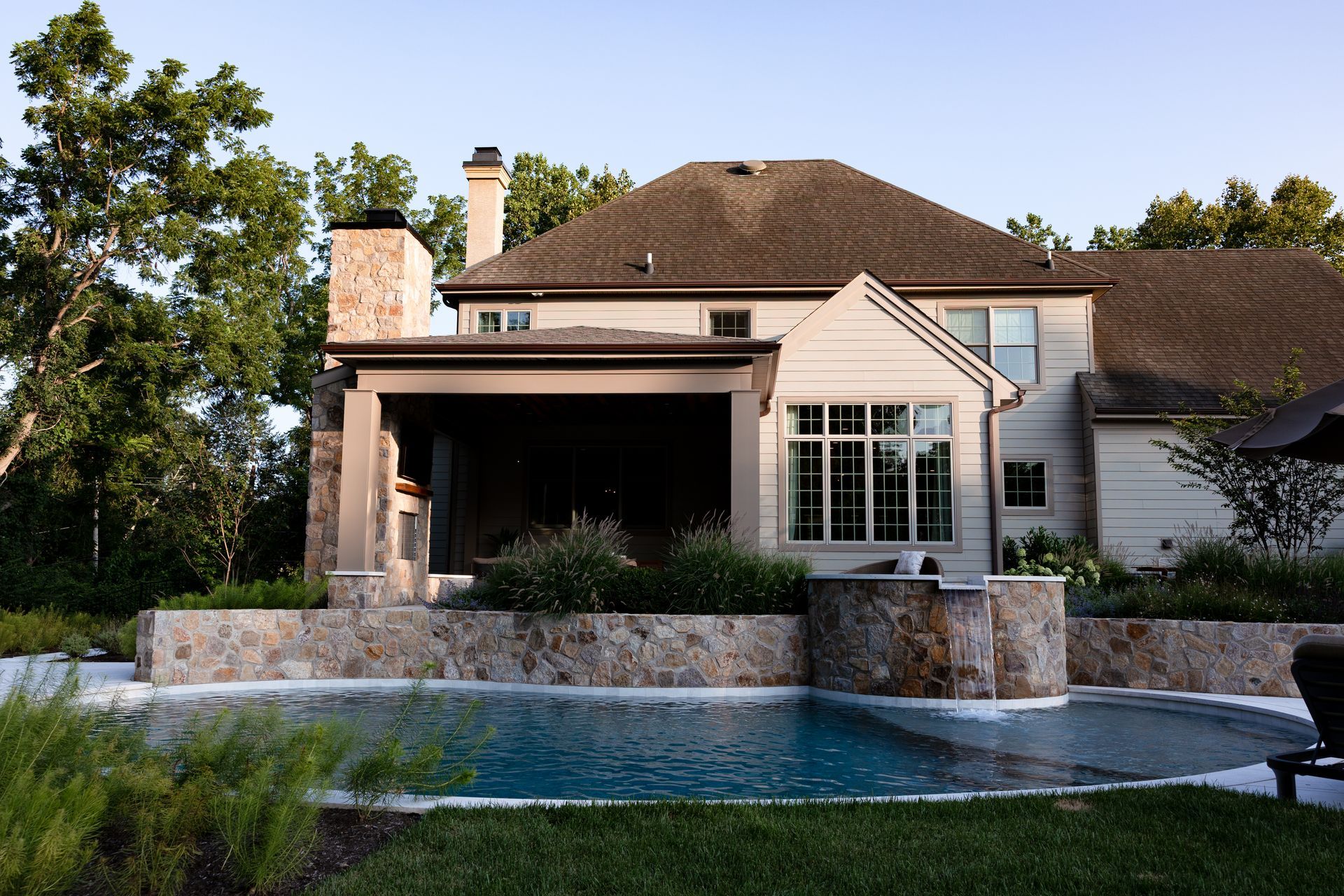 Backyard pool with a house, featuring stone wall, covered patio, and lush greenery.