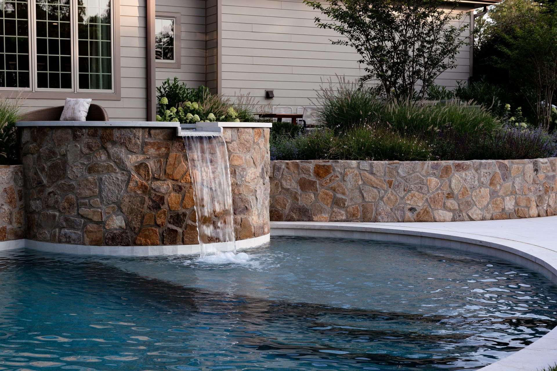 Swimming pool with stone waterfall feature, built-in planters, and a house in the background.