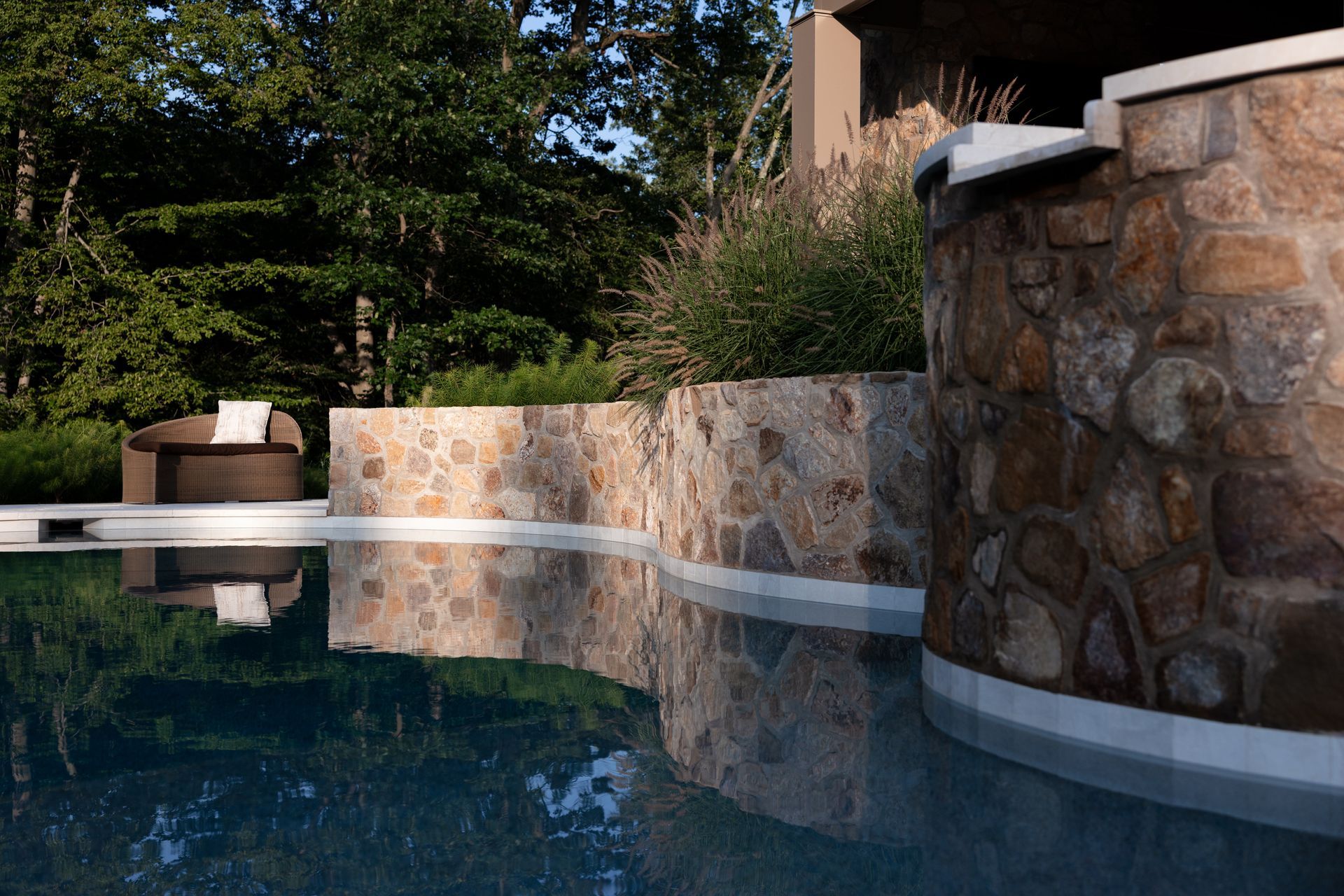 Swimming pool with stone walls reflecting in the water, a lounge chair in the background, trees.