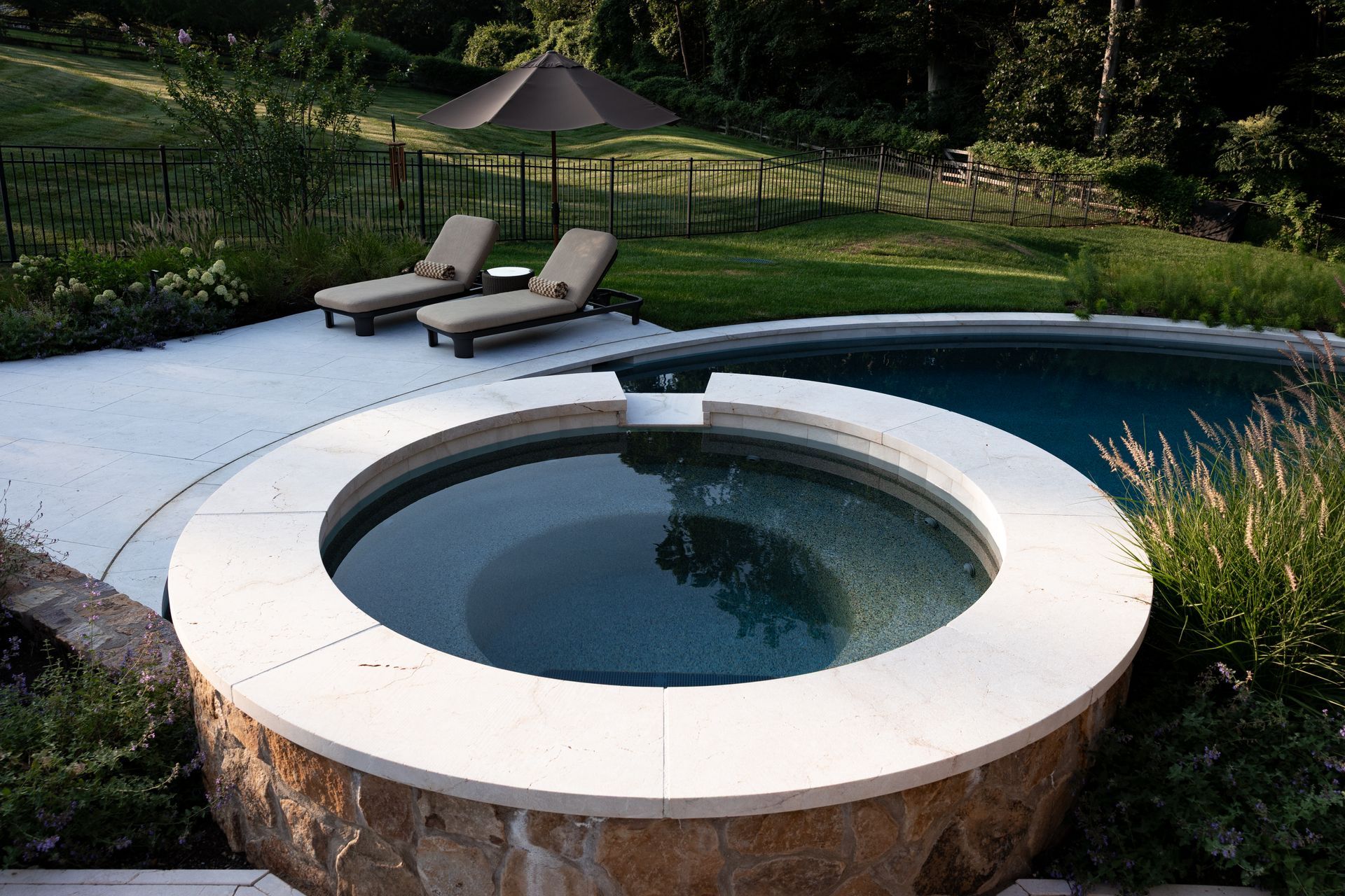 Outdoor spa and pool with lounge chairs and umbrella in a landscaped setting.
