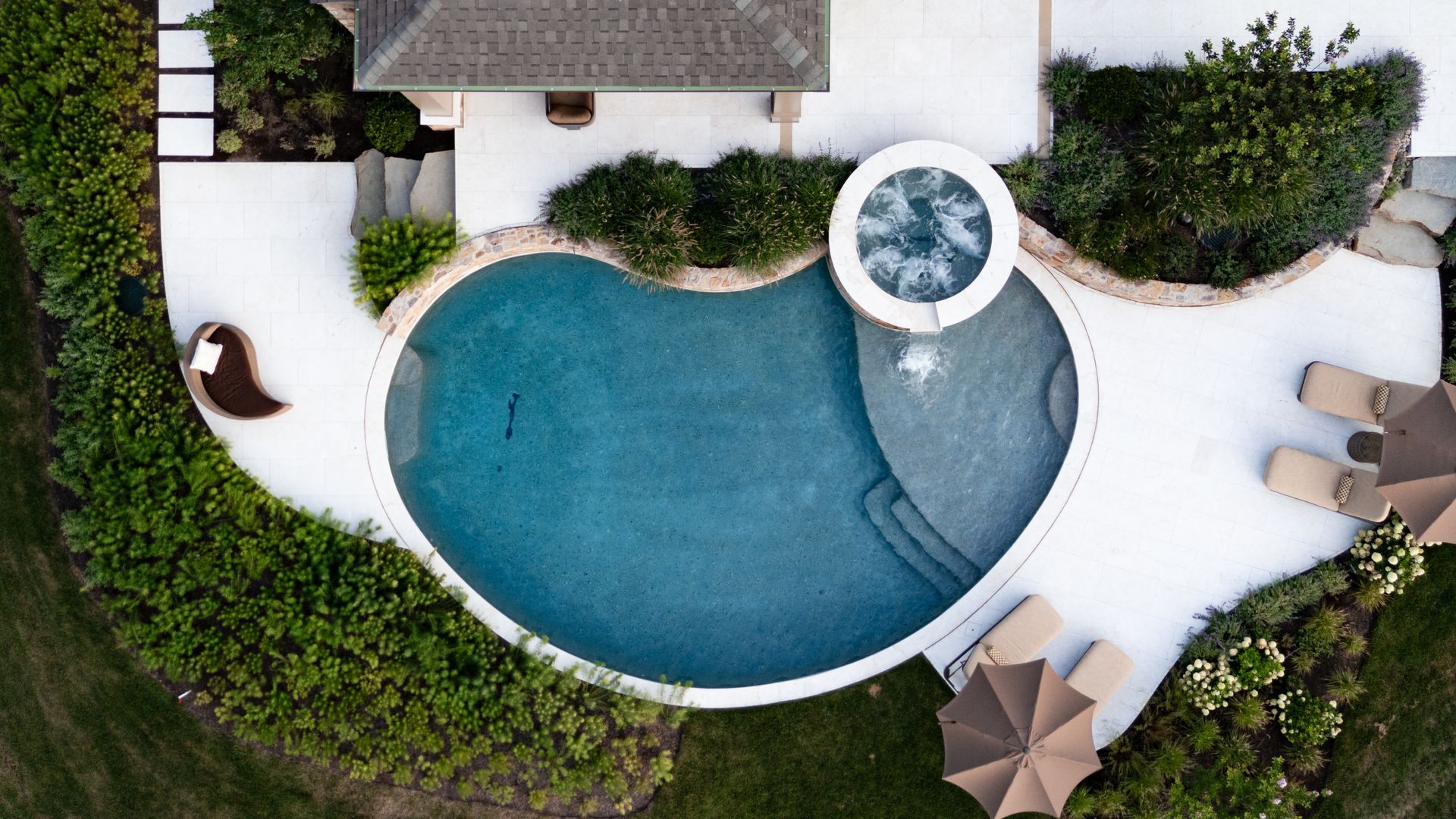 Aerial view of a swimming pool with a connected jacuzzi, surrounded by white concrete and landscaping.