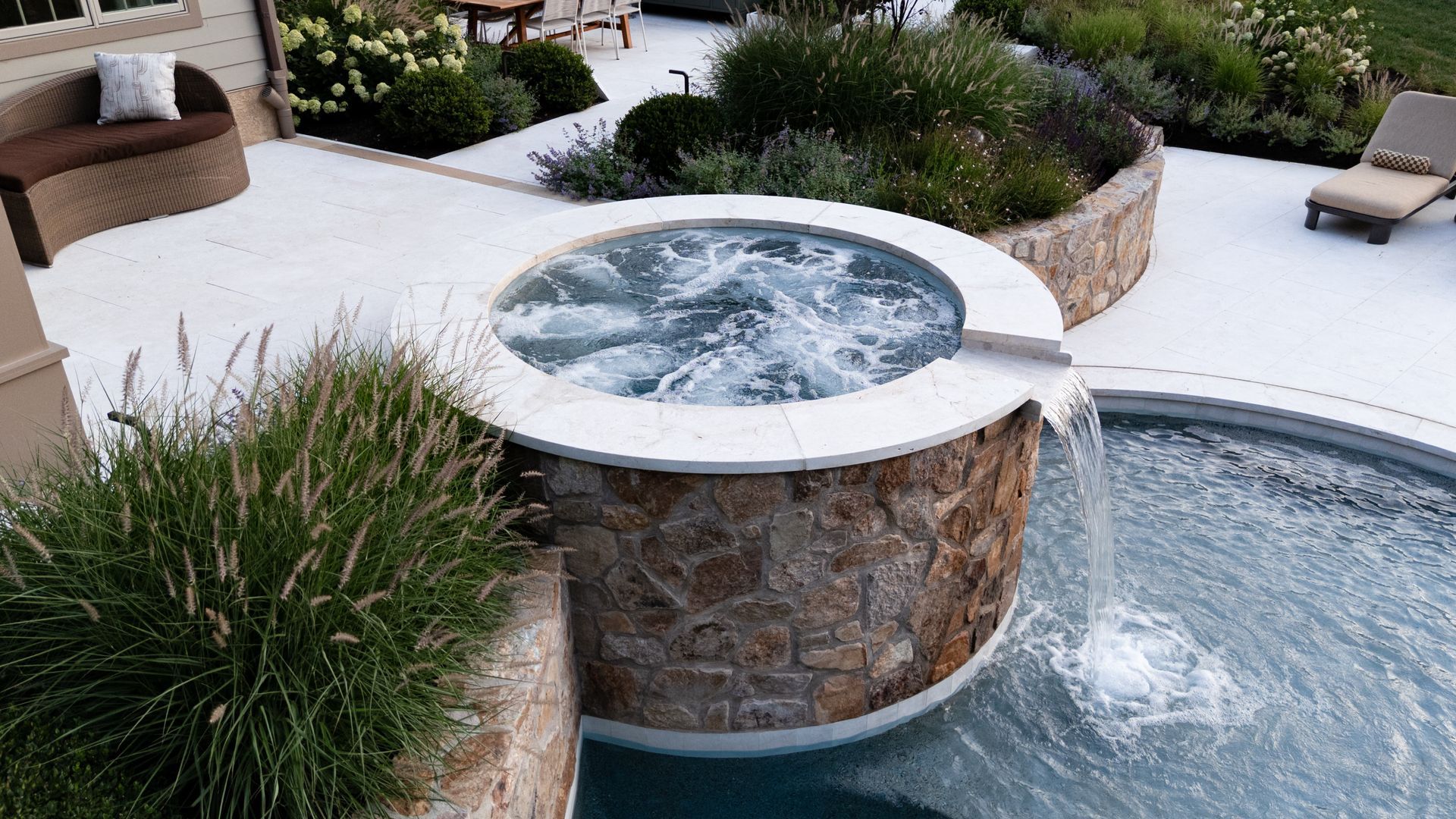 Hot tub overflowing into a pool. Stone-lined with light-colored patio and landscaping.