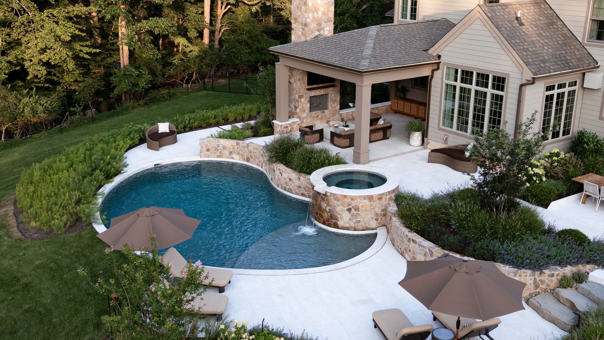 Luxury backyard with pool, spa, patio, and outdoor kitchen. Beige stone, tan umbrellas, lush greenery.