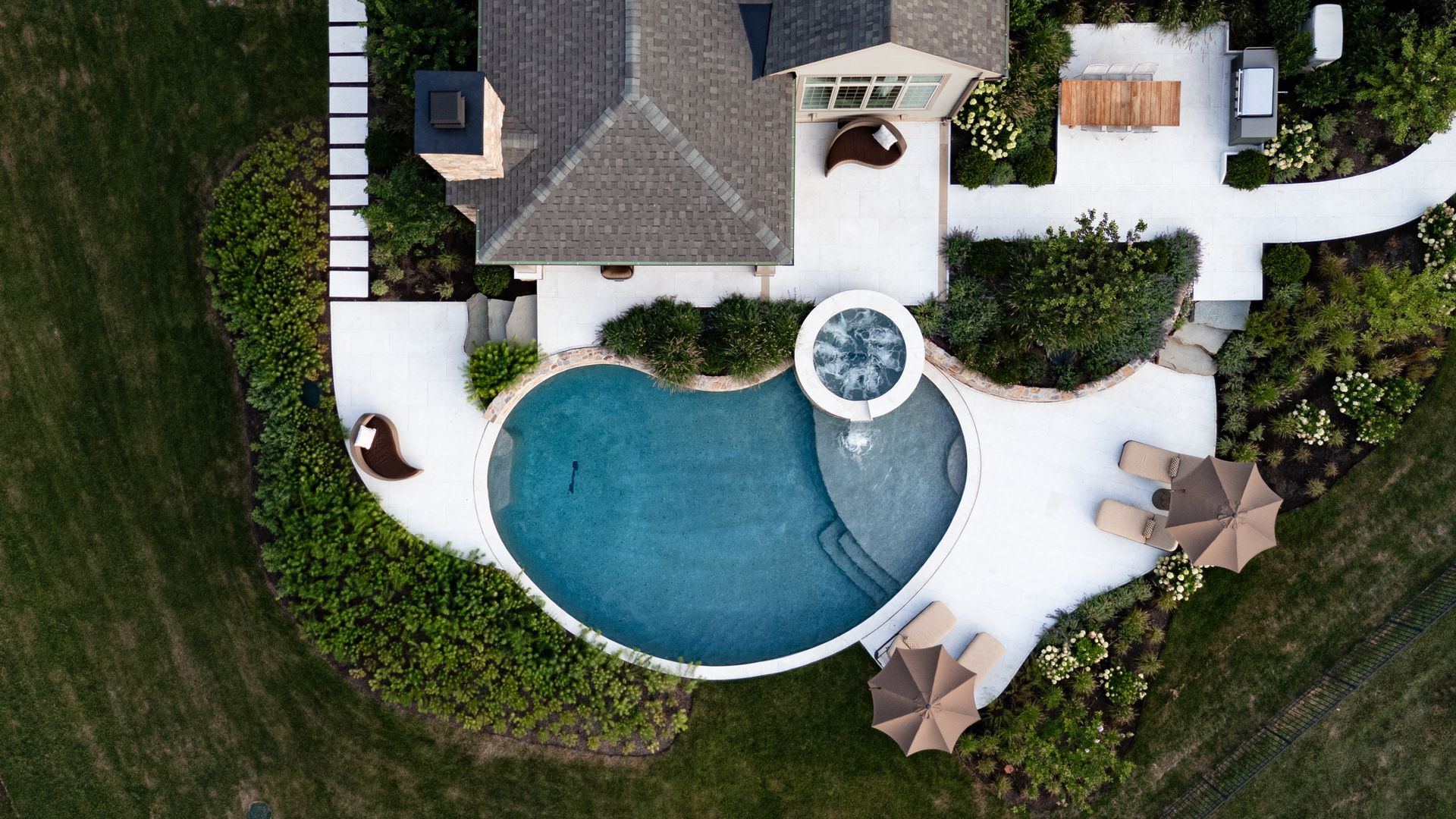 Overhead view of a house with a pool and patio. Green lawn surrounds a white patio, pool, and seating areas.