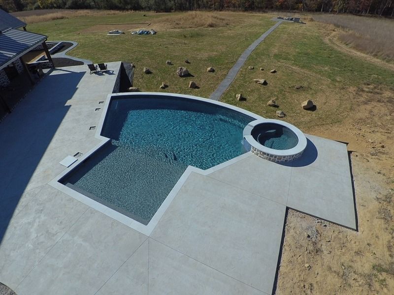 Aerial view of a swimming pool and hot tub surrounded by concrete patio, with a grassy yard and trees in the distance.