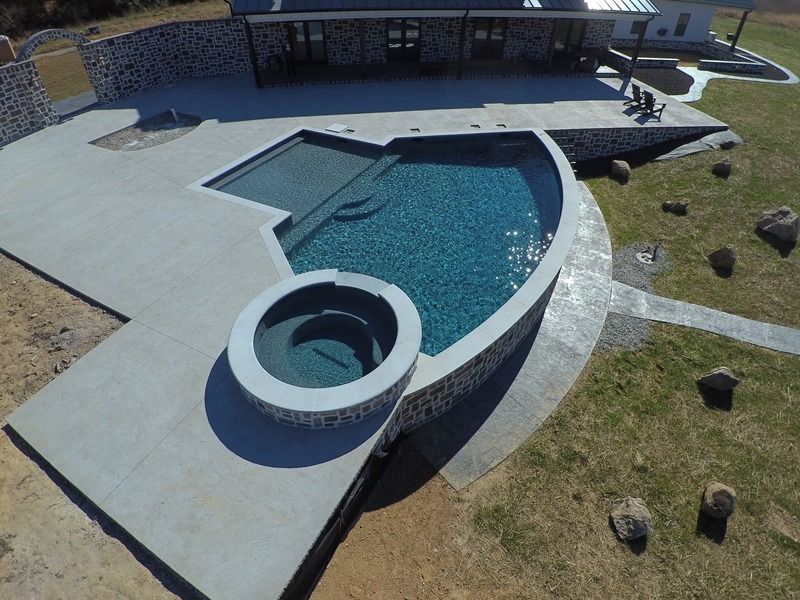 Aerial view of a custom pool and spa with stone accents surrounded by a concrete patio and green lawn.