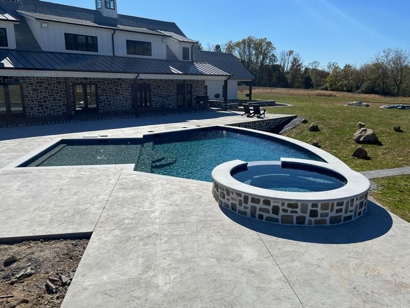 Pool and spa with stone accents beside a large white house on a grassy property.