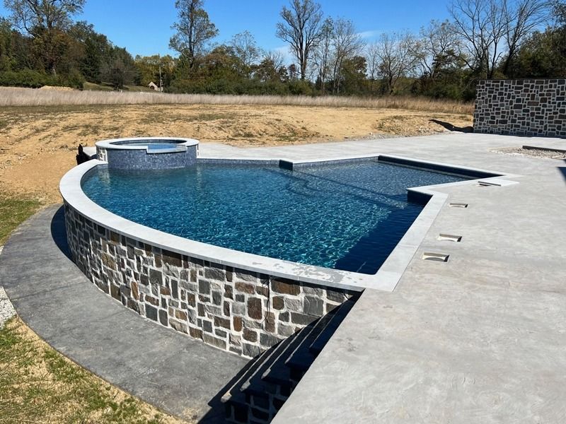 A newly constructed swimming pool with a hot tub, surrounded by a stone wall and concrete patio, outdoors.