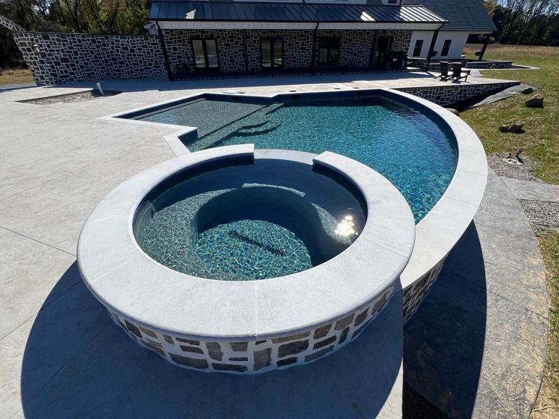 Pool with a curved edge and a circular spa. Blue water, gray stone deck, and a house in the background.