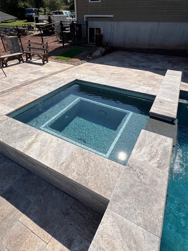 Square hot tub integrated into a pool with stone surround; exterior setting.
