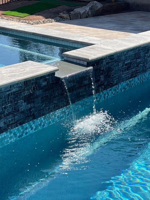 Water cascades from a dark, tiled wall into a swimming pool. Sunlight reflects on the clear blue water.