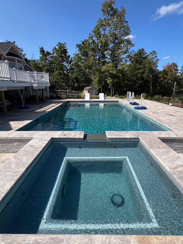 Swimming pool with an attached spa, light-colored stone surround, blue water, and a house in the background under a blue sky.