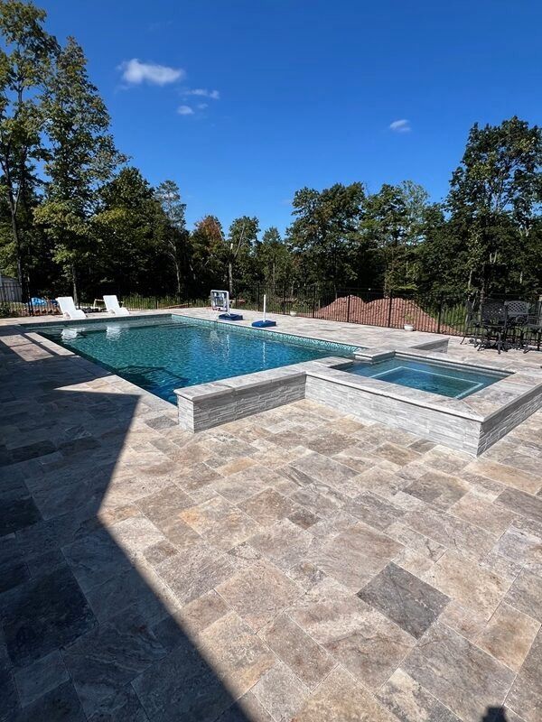 Pool and hot tub with stone surround, blue water, blue sky, and trees in the background.