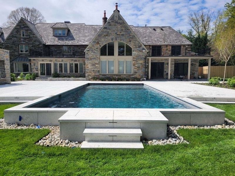 Backyard with rectangular pool and stone house, gray patio, green grass, and small rocks.