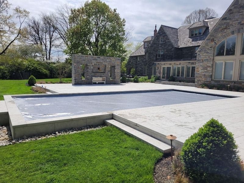 Pool with a blue cover, stone patio and landscaping, stone house in background.