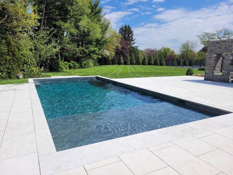 Rectangular swimming pool with light gray tile surround, set in a sunny, grassy backyard.