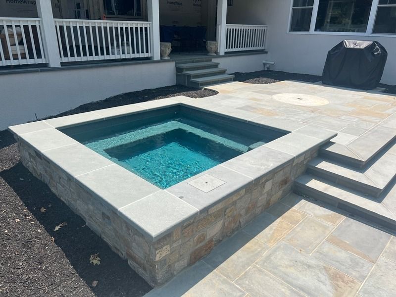 Square spa with teal water, stone surround, steps, and patio.