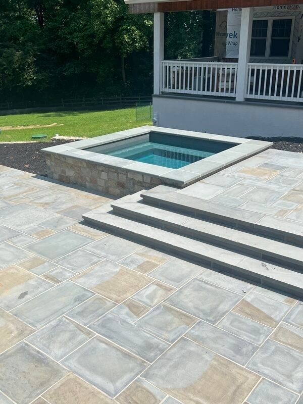Square hot tub built into stone patio with steps, near a house with a porch.