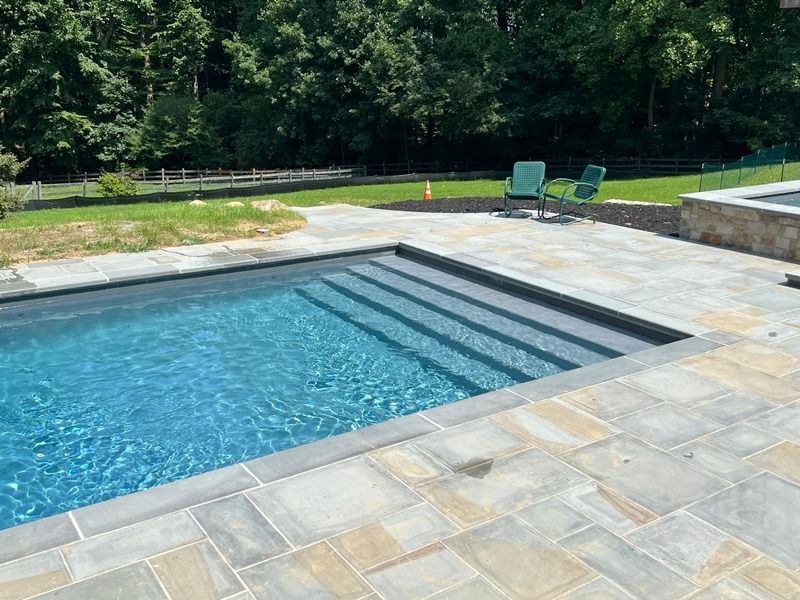 Pool with built-in steps, surrounded by stone patio. Two chairs sit on the lawn. Trees in the background.