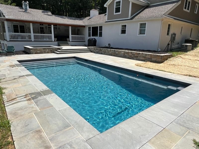 Rectangular pool with blue water and steps, surrounded by stone patio, next to a house with a porch.