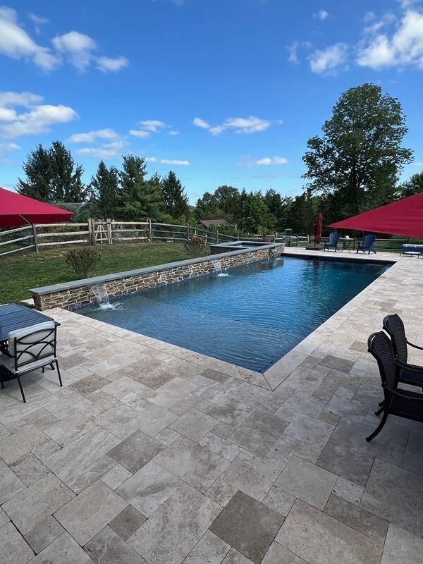 A rectangular swimming pool with water fountains, surrounded by stone patio and red umbrellas, under a blue sky.