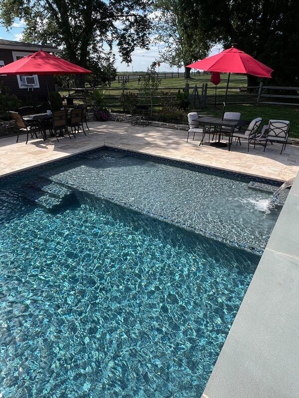 Swimming pool with red umbrellas, tables, and chairs on a stone patio, with a grassy area and trees.