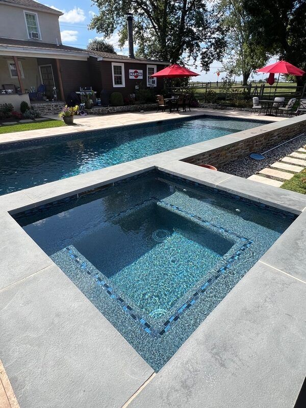 Backyard with a dark blue pool and hot tub, surrounded by stone. Red umbrellas and a house are visible.