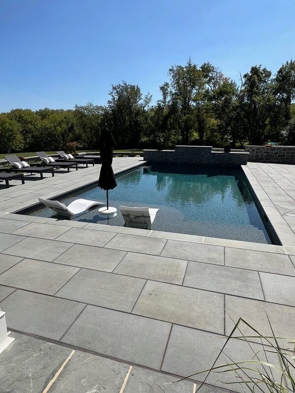 Pool with lounge chair and umbrella on a sunny day. Gray stone patio, trees in background.