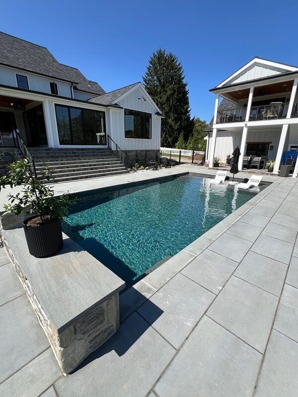 Rectangular turquoise pool surrounded by gray stone patio, adjacent to a white house with a two-story covered porch.