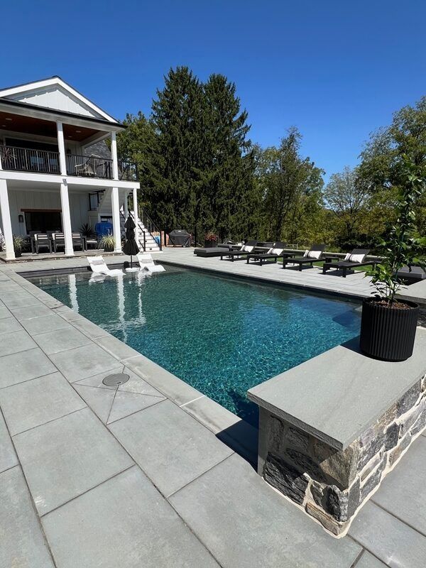 Pool with clear blue water and stone patio, adjacent to a white house with a balcony.