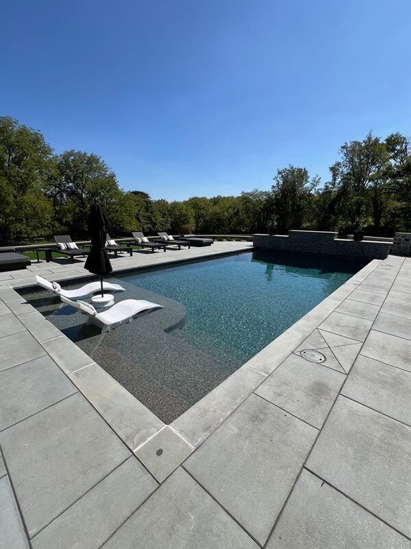 Pool with lounge chairs and umbrella on a sunny day. Large stone patio and trees in the background.