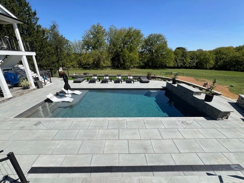 Pool with lounge chairs and stone patio on a sunny day. Green lawn and trees in the background.