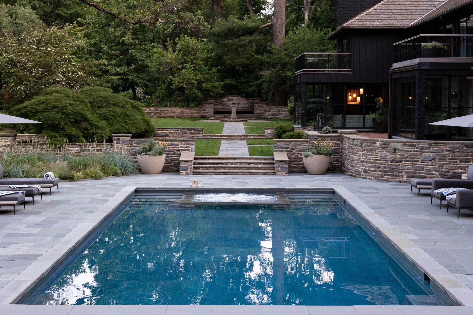 Rectangular pool with gray stone patio, overlooking a tiered yard and black house with a thatched roof.