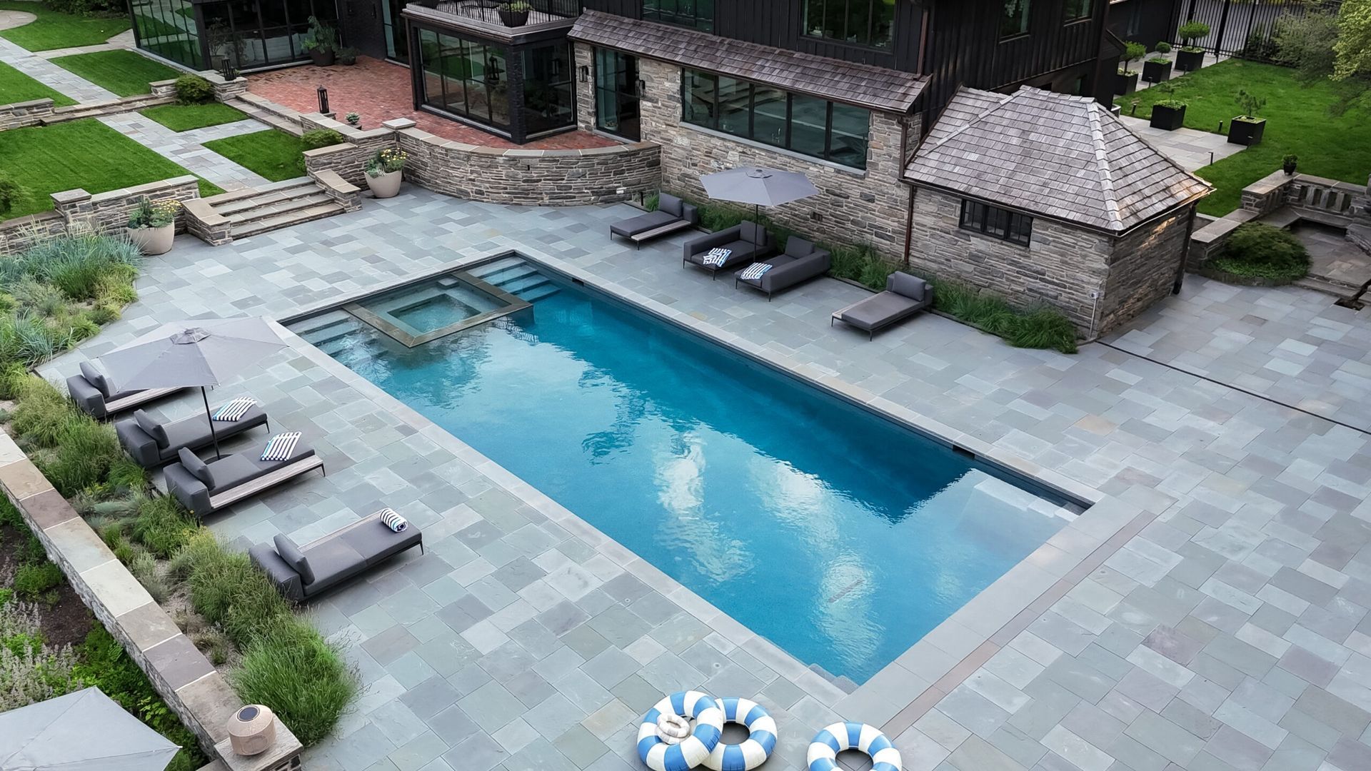 Overhead view of a luxurious outdoor pool area with a dark house, stone patio, and lounge chairs.