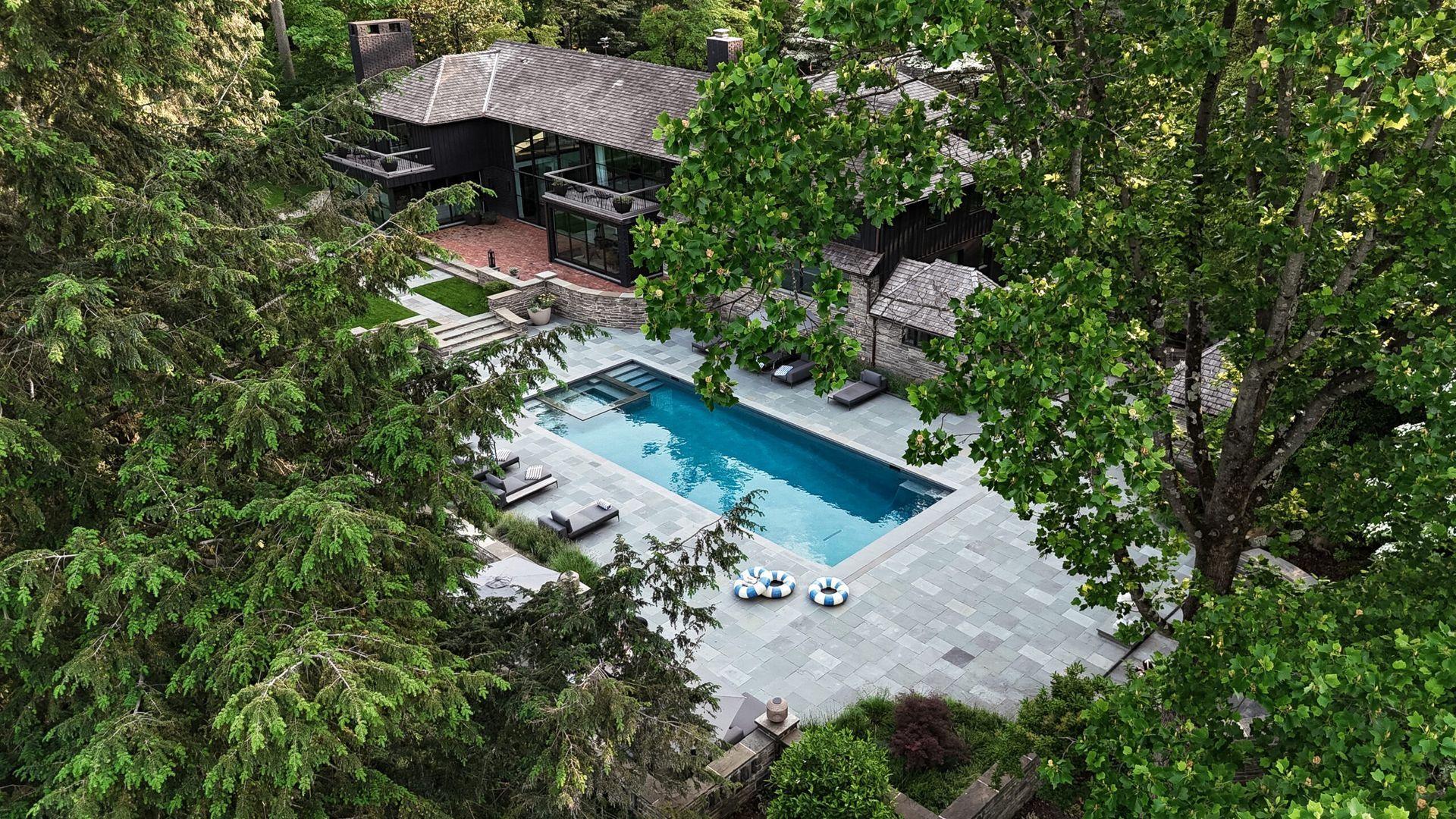 Aerial view of a home with a rectangular pool surrounded by trees and a stone patio.