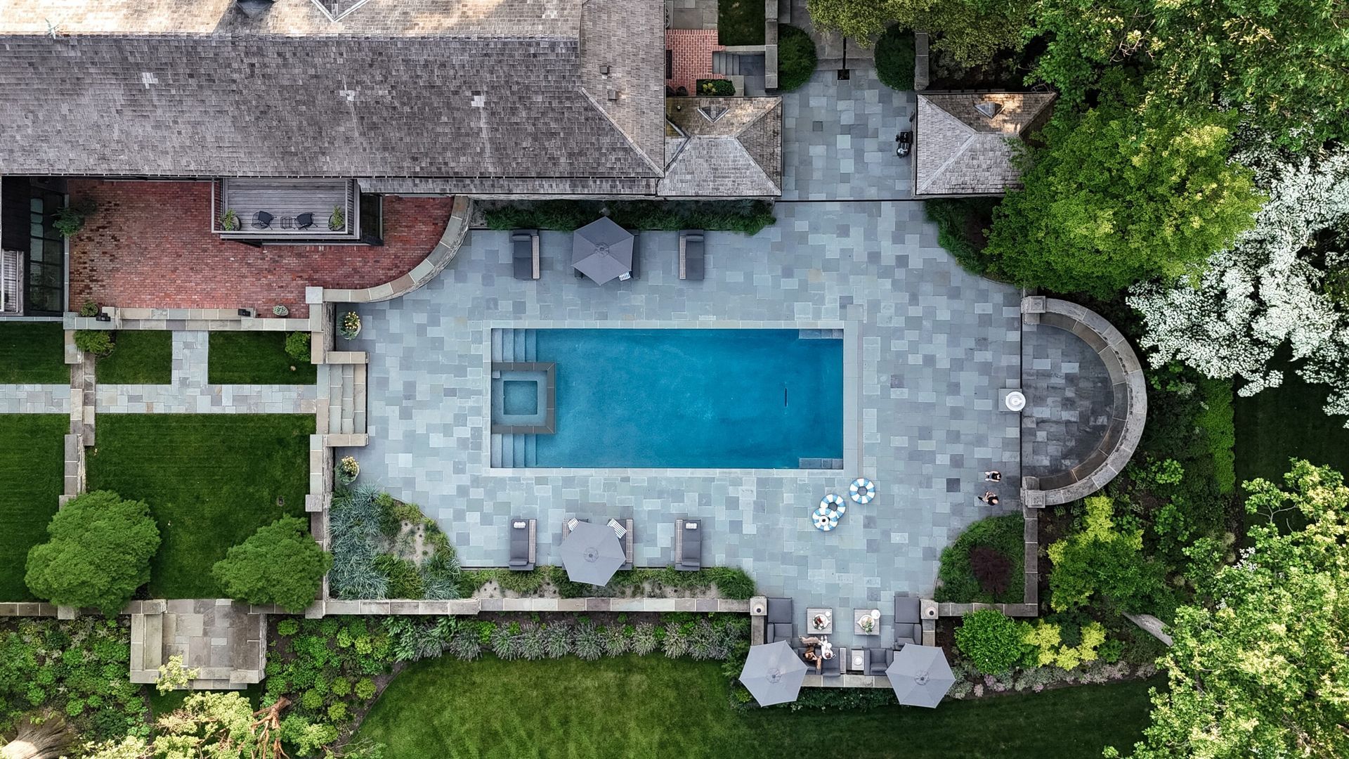 Overhead view of a rectangular pool surrounded by a stone patio, lounge chairs, and manicured landscaping.