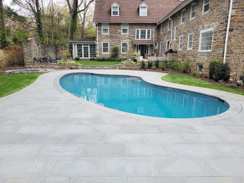 Pool with light blue water and stone patio, in front of a multi-story stone house with a lush green lawn.