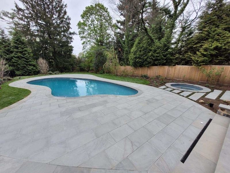 Backyard with a curvy pool, gray paving, and a hot tub. Trees surround the area.