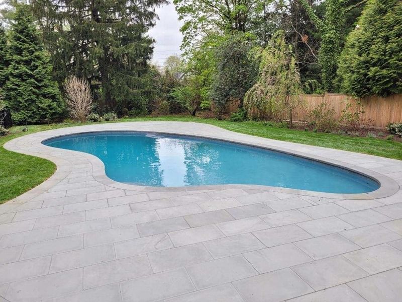 A freeform swimming pool surrounded by gray stone patio and lush green grass; trees in the background.