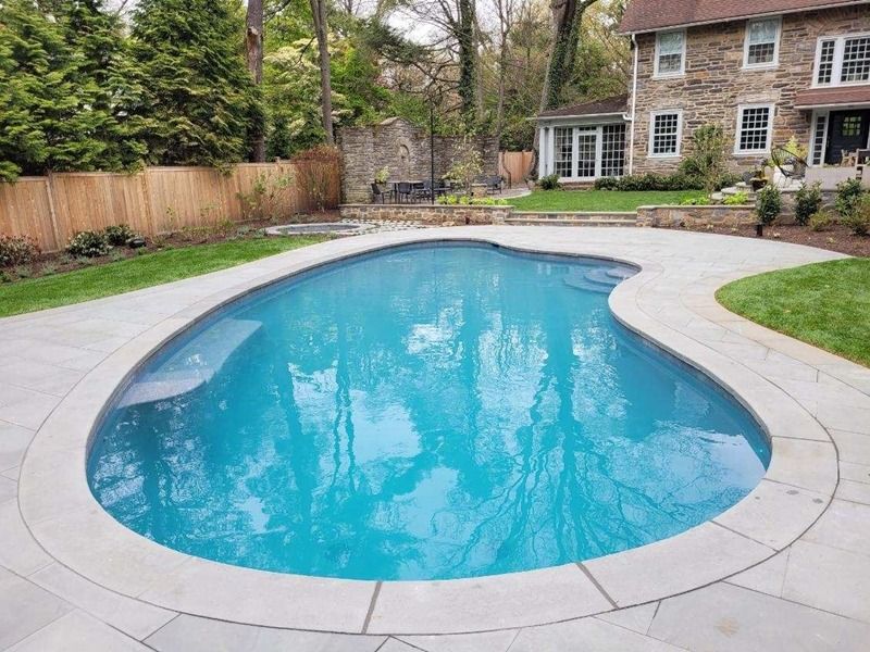 Swimming pool surrounded by stone patio and green lawn, with a brick house in the background.