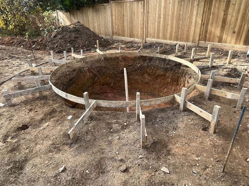 Circular hole in ground with wooden framework, likely for a pool or foundation; construction site.