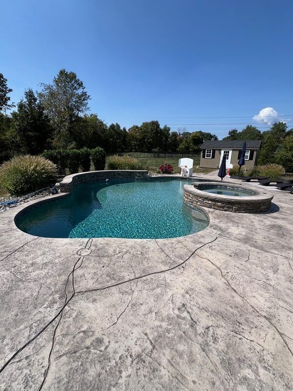 Pool with turquoise water and a hot tub set in a concrete patio, surrounded by greenery under a blue sky.